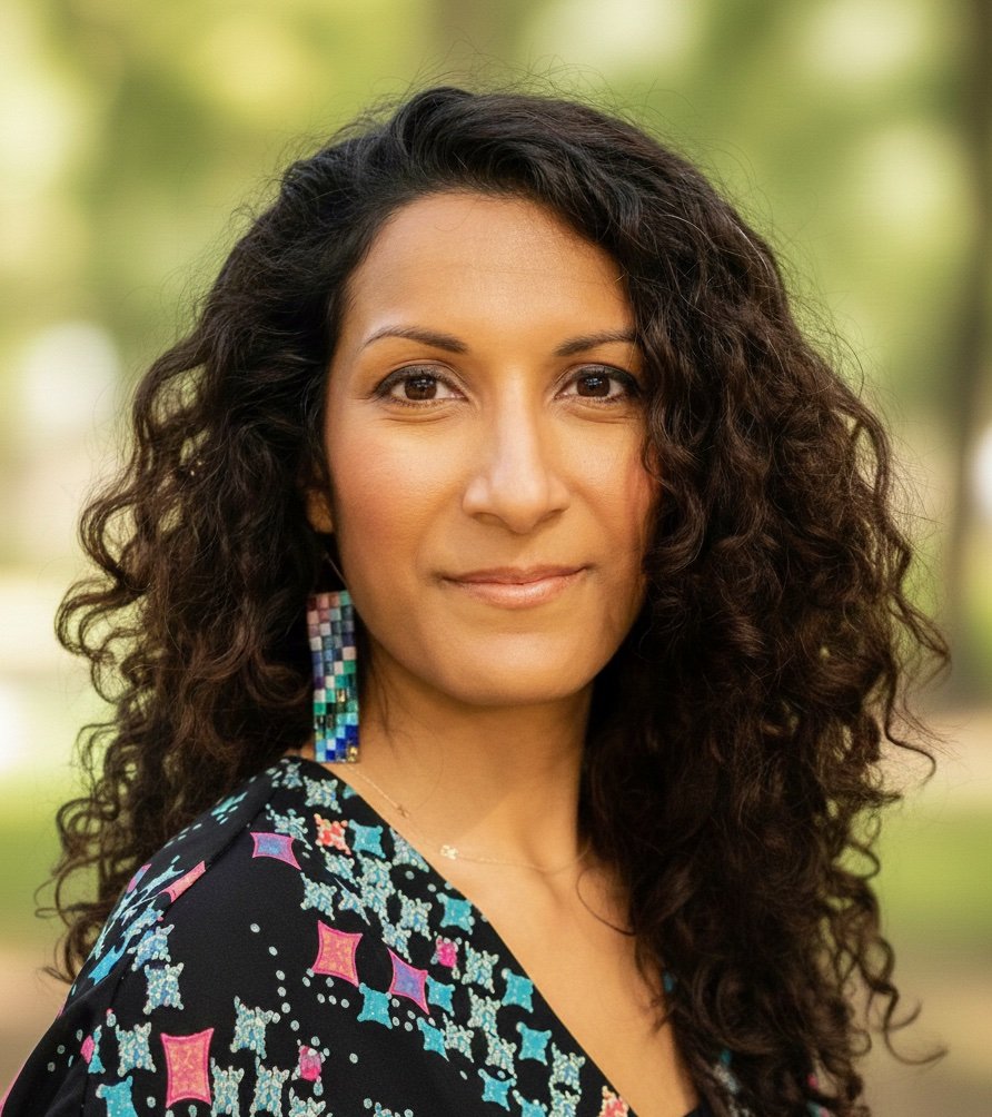 Headshot of Dr. Roopa Kalyanaraman Marcello. She is a woman with curly dark brown hair, wearing patterned earrings and a colorful patterned dress. In this photo, she is outdoors with a blurred leafy background.