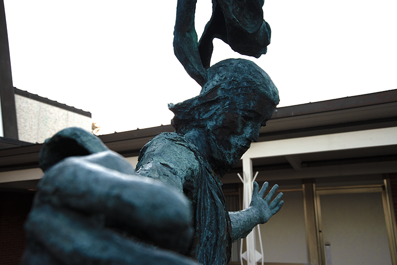 Close-up of a bronze statue of Jesus Christ His arm extended outward, set against a background of a building and cloudy sky.
