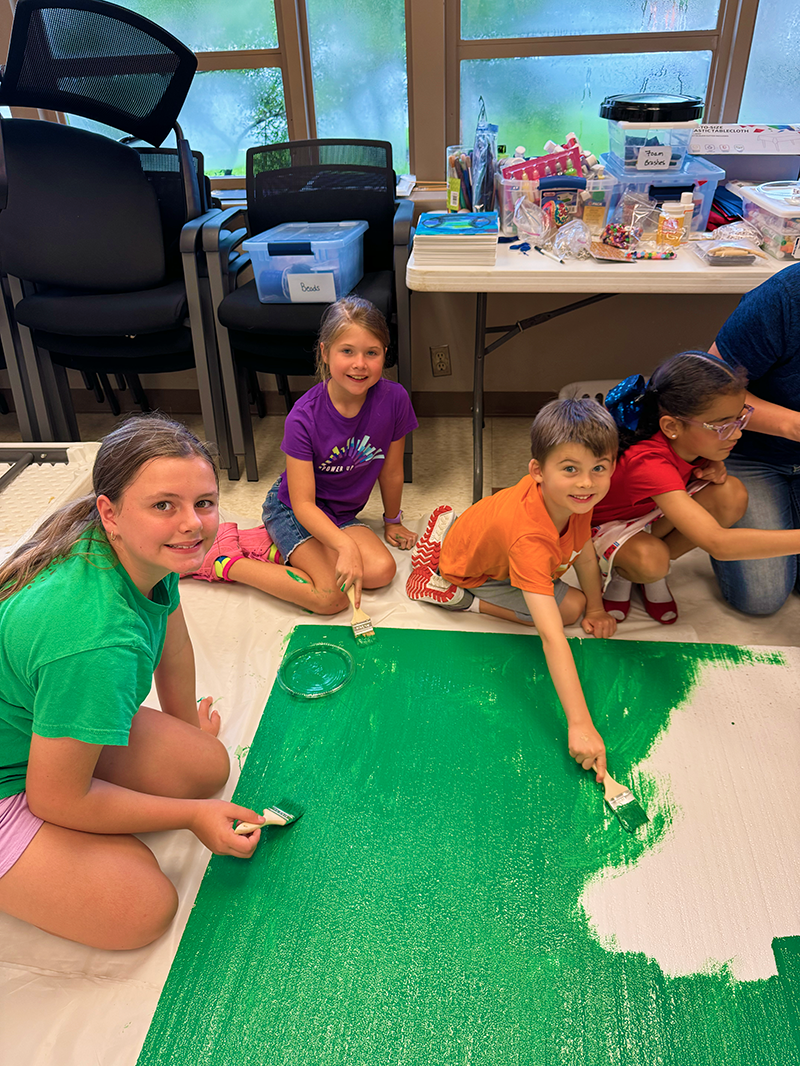 Children painting a large white canvas with green paint, sitting on the floor in a room with art supplies and chairs.