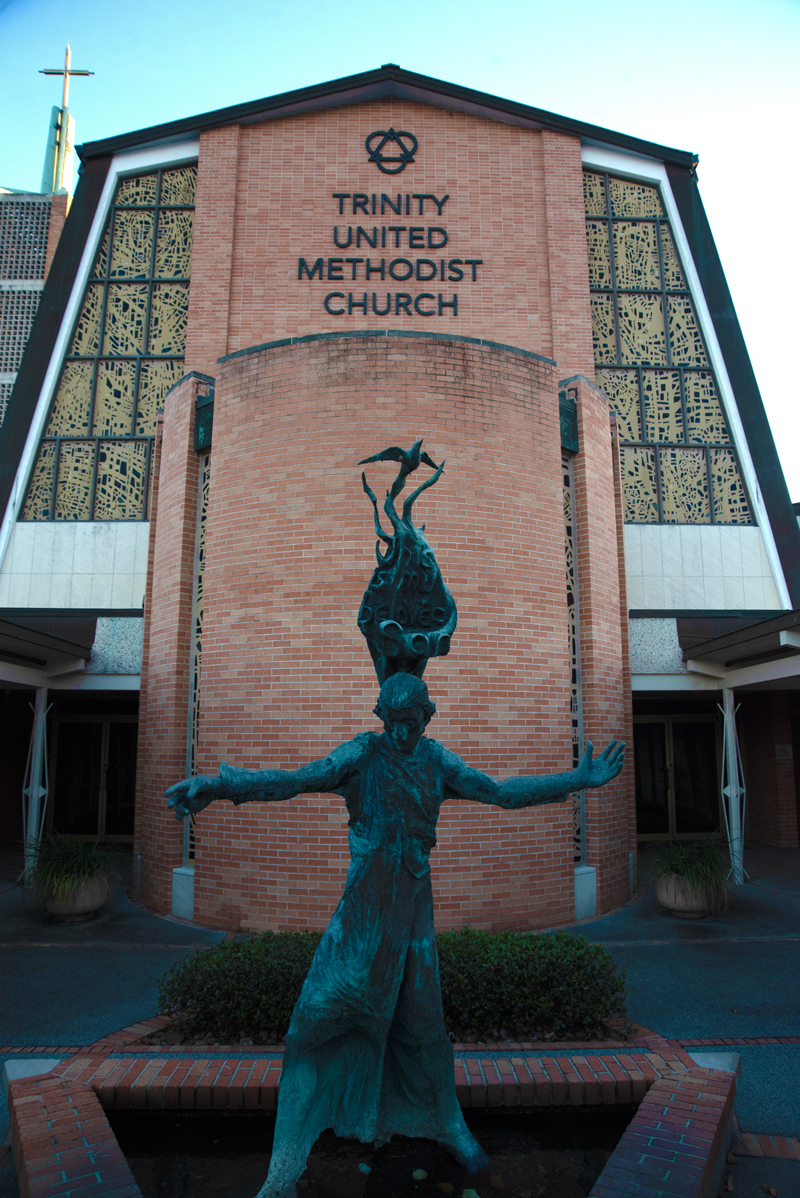 The front of Trinity United Methodist Church featuring a brick facade, a prominent sculpture of Jesus Christ with a dove alighting the top of the statue, and stained-glass windows.