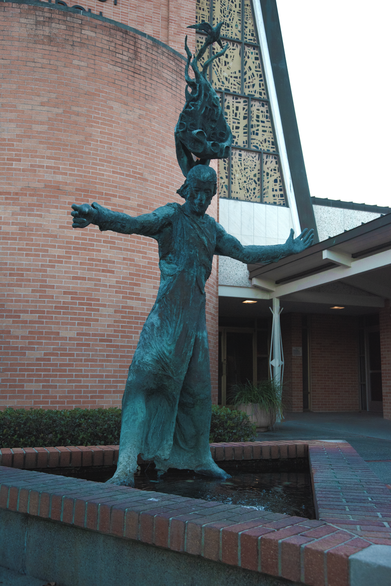 A bronze sculpture of Jesus Christ standing in a water feature with his arms outstretched. The sculpture is outside a brick building with decorative metalwork on a window.