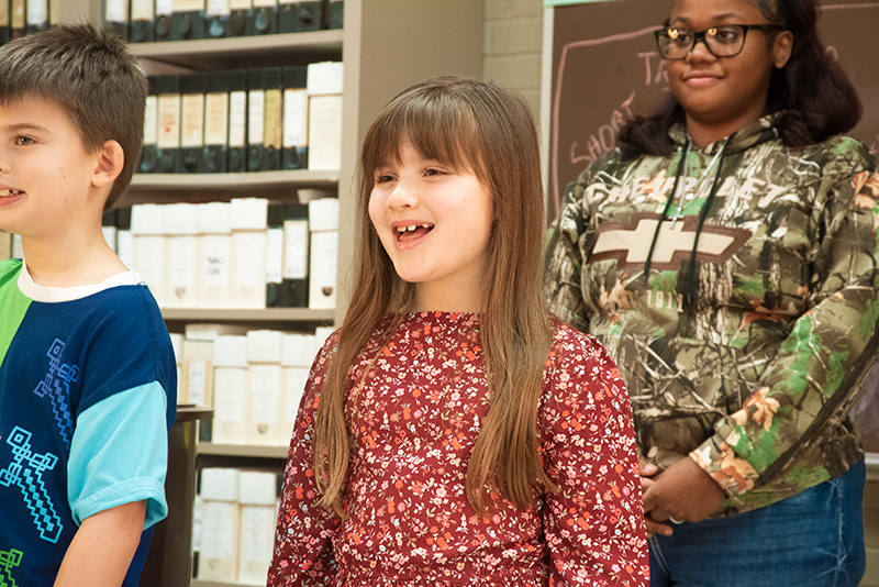 Three children standing and singing indoors, with shelves of music in the background.