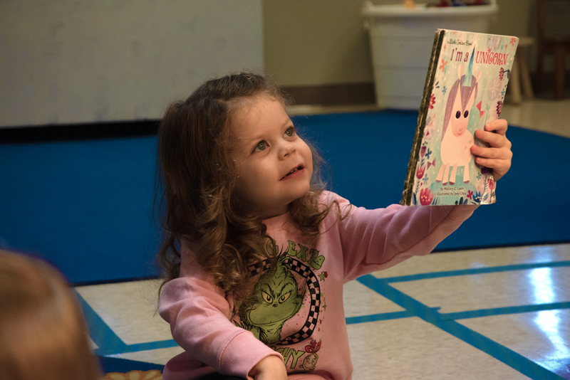 A young girl with curly brown hair is holding a colorful children's book titled 'I'm a Unicorn' and looking at it with curiosity. She is sitting on a gymnasium floor.