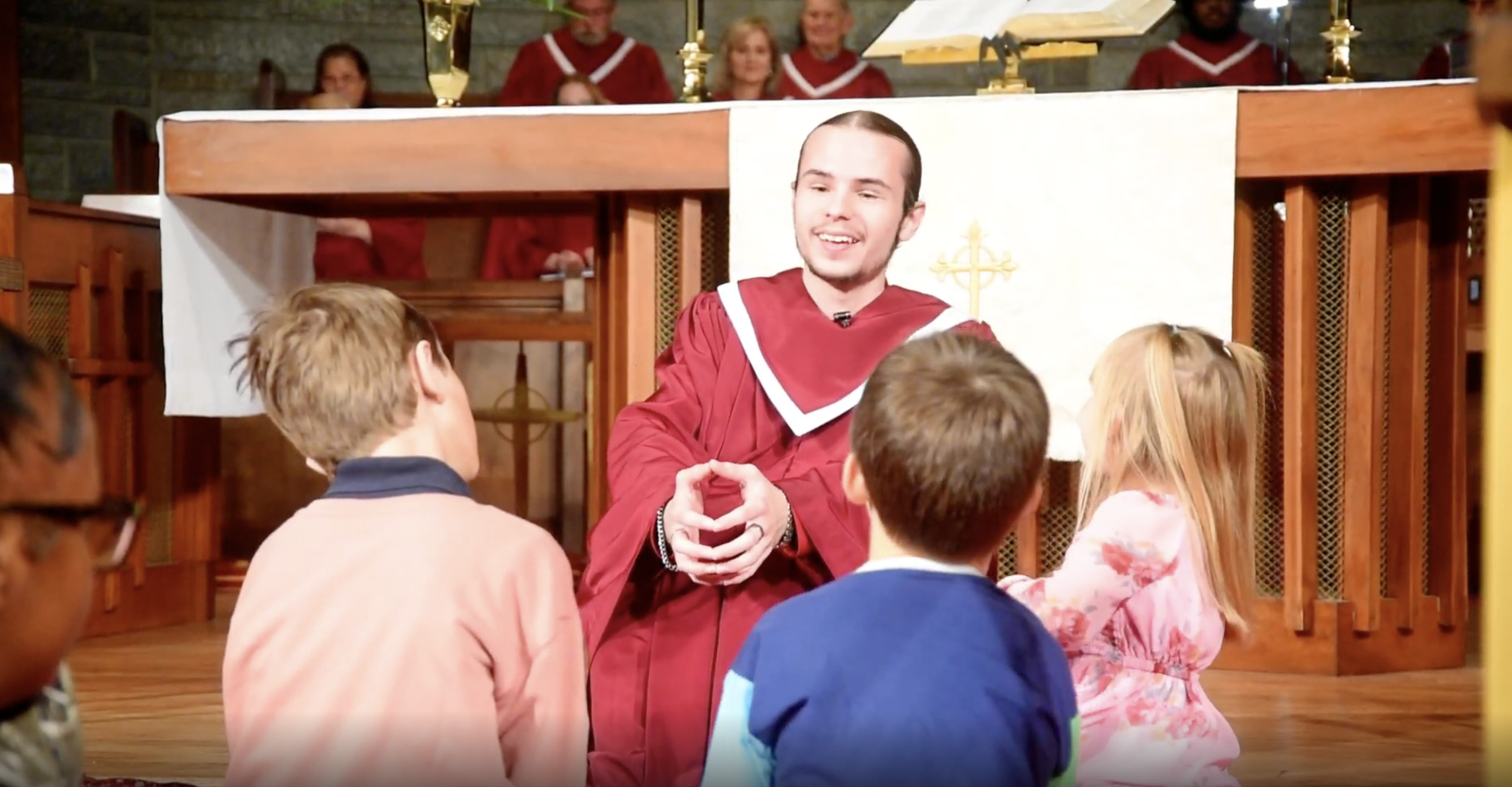 A church scene with a preacher in a maroon robe sitting on the floor, talking to children sitting in front of him, during a children's church service.
