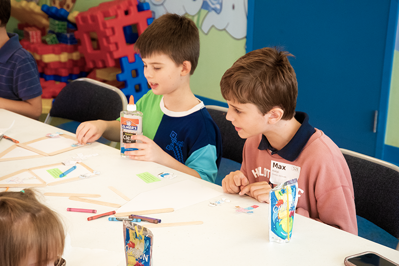 Two boys sitting at a table with craft supplies, working on a project, in a colorful room with building blocks in the background.