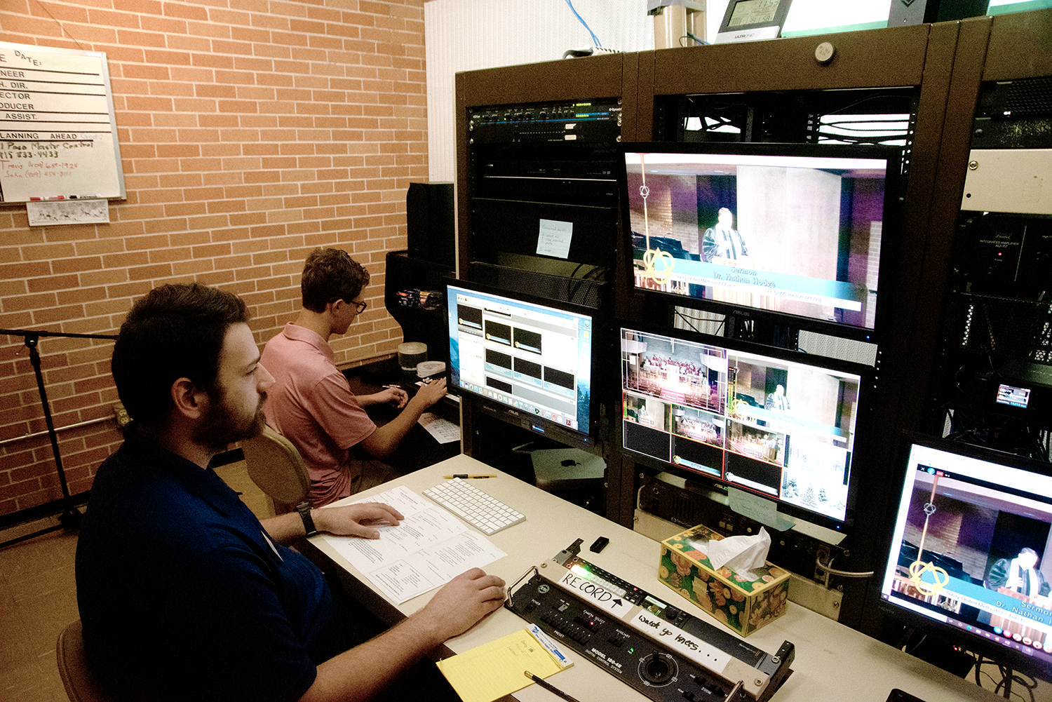 Two men working at a video control station with multiple monitors displaying video feeds and editing software. The man in the foreground is wearing a dark shirt and working with papers, while the man in the background is wearing glasses and a pink shirt, operating a computer. The room has a brick wall and a whiteboard on the wall.