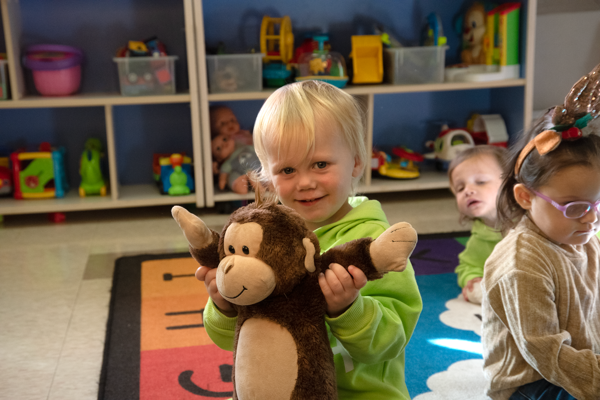 A young boy with blonde hair wearing a green hoodie is holding a plush monkey toy and smiling at the camera. In the background, two other children are sitting on a colorful rug in a playroom with shelves filled with toys and stuffed animals.