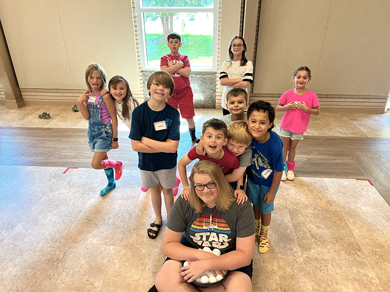 A group of children and one adult man posing together inside a room with a large window in the background. The children are smiling and making playful gestures.