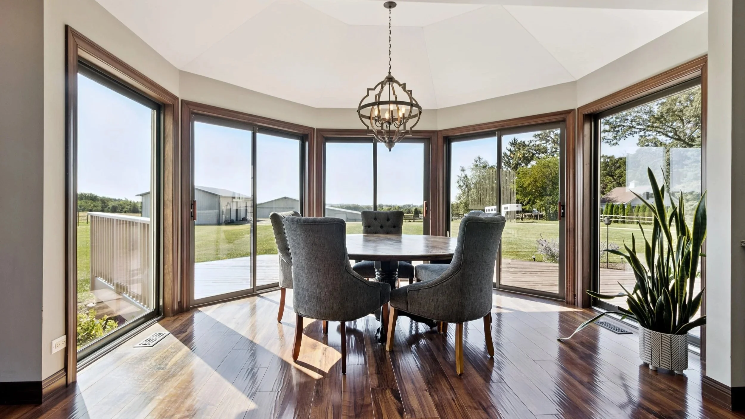 Sunlit dining area with a round table and six upholstered chairs in front of large glass sliding doors that open to a wooden deck and expansive green yard outside.