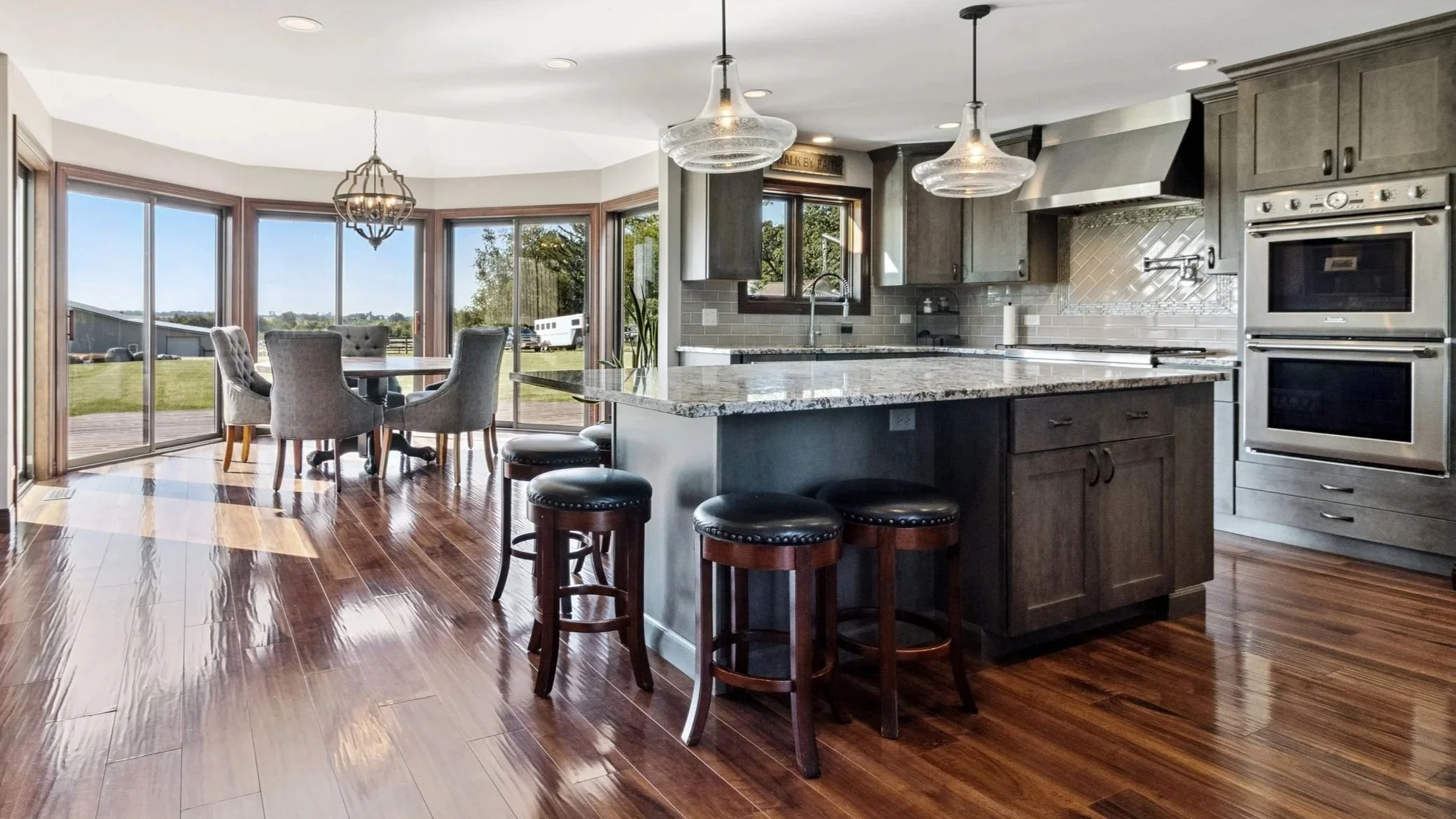 Open-concept kitchen with gray cabinets, granite countertops, and stainless steel appliances, adjacent to a dining area with a round table and six chairs, large windows and a sliding glass door overlooking a grassy yard and blue sky.