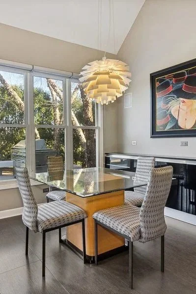 A dining room with a glass-top table, four patterned upholstered chairs, large windows showing trees, a hanging modern pendant light, a colorful abstract painting, and a black piano.