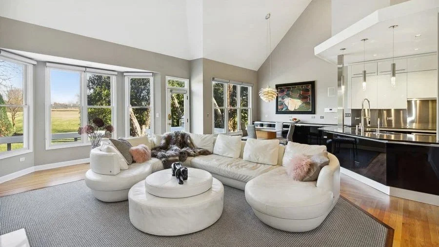 Bright living room with large windows, white curved sectional sofa with pink and animal print pillows, circular ottoman with a black decorative object, and an open kitchen with stainless steel appliances and black cabinetry.