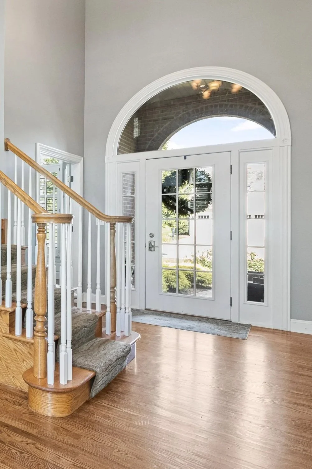 Residential entryway with wooden staircase, gray walls, and a glass-paneled door with an arched window above, letting in natural light.