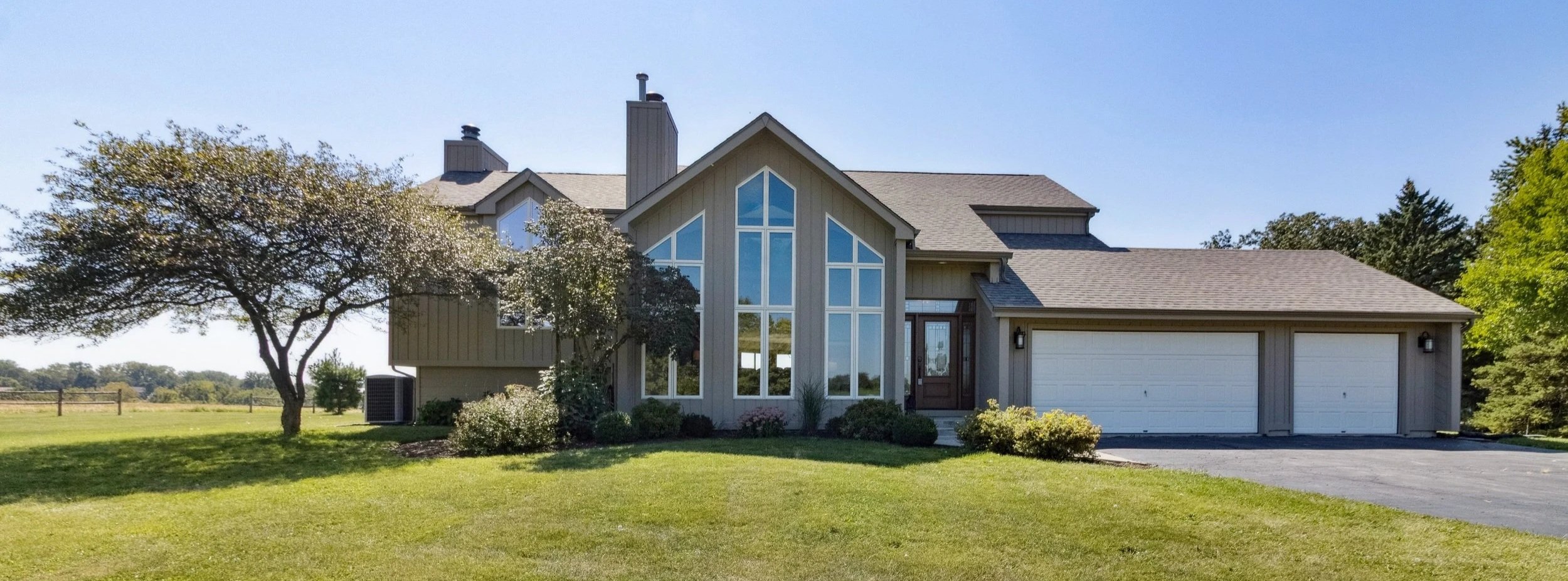 A modern two-story house with large windows, a gray exterior, a two-car garage, surrounded by a green lawn and trees under a clear blue sky.
