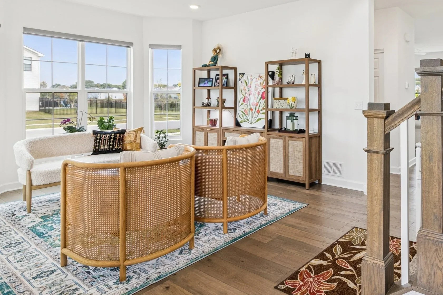 Bright living room with large windows, white walls, a patterned area rug, a white textured sofa, two rattan chairs, and a wooden bookshelf with decorative items and artwork.