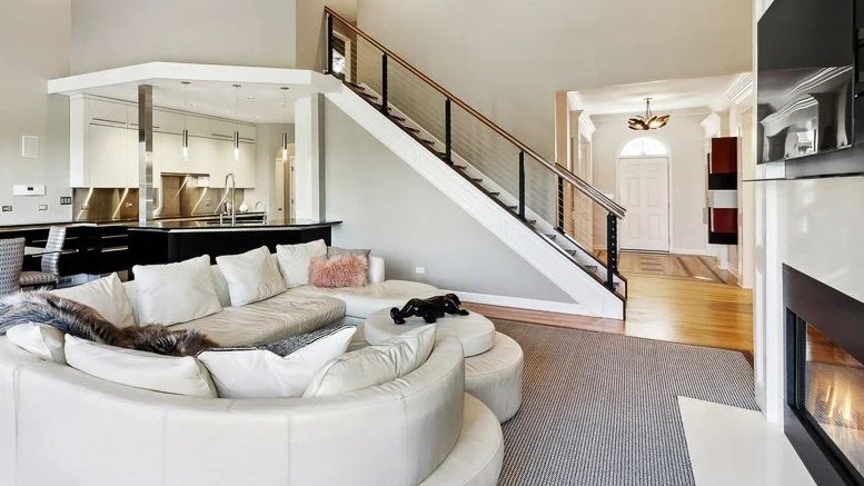 Living room with a white curved sofa, some pillows, and a staircase with black railing leading to the upper floor, open to a kitchen area.