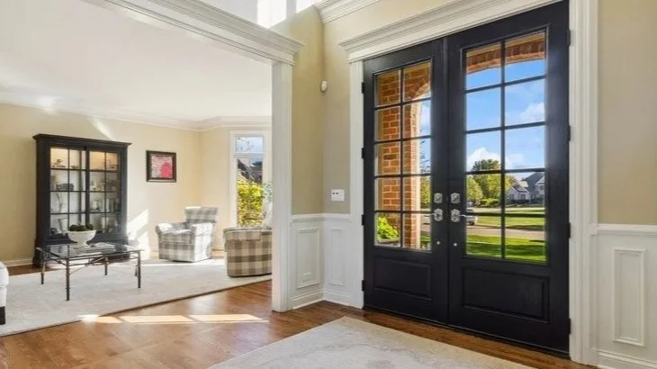 Interior view of a home's entryway with black double glass doors leading outside to a green lawn, and a living room with beige walls and dark furniture.