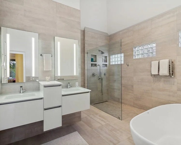 Modern bathroom with dual sinks, large mirror, walk-in shower with glass door, and a bathtub. Light-colored tiles on walls and floor, towel rack with two towels, small shelves in shower, and glass blocks for natural light.