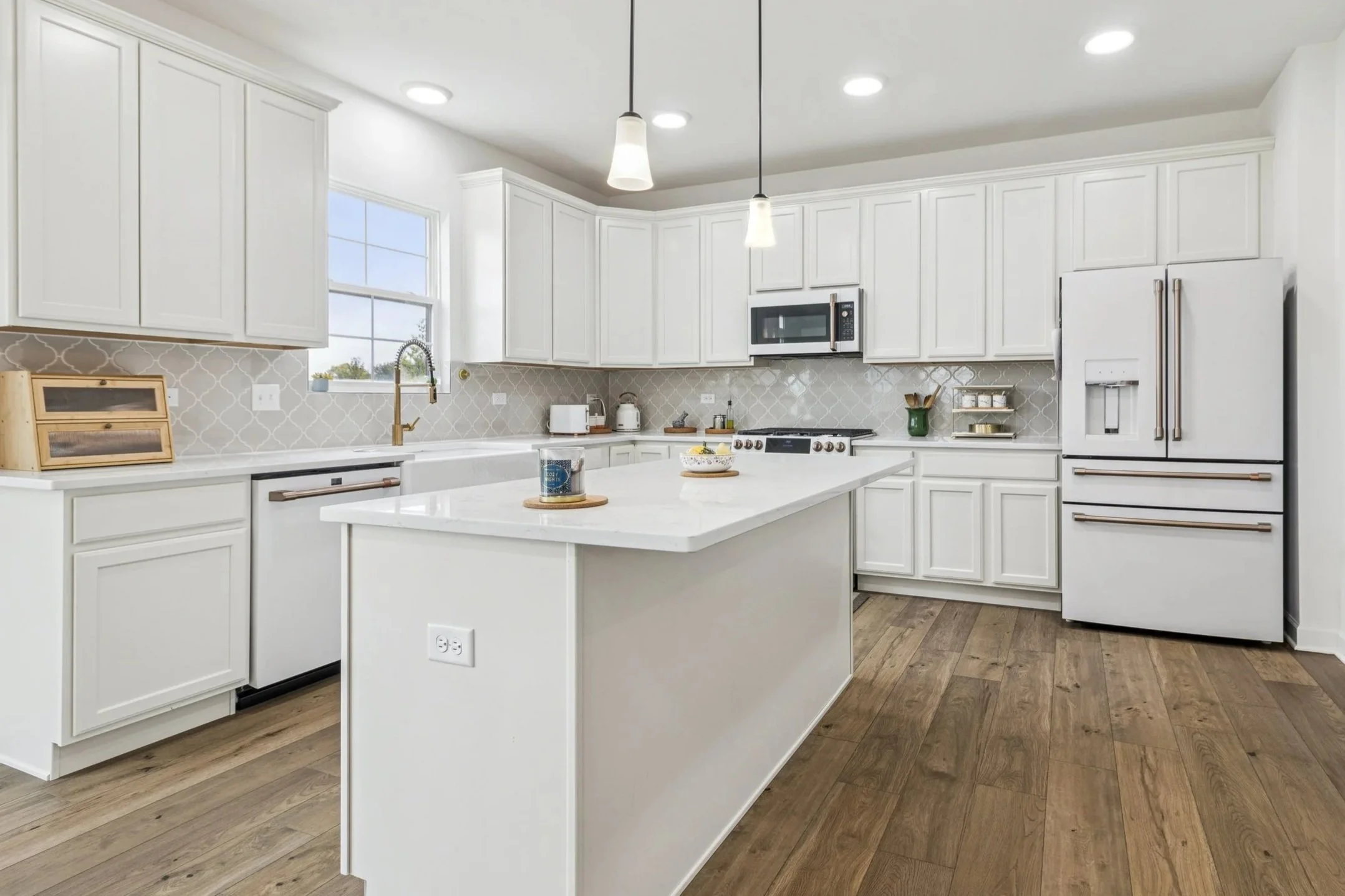 Modern white kitchen with wooden floors, island, and white cabinetry, featuring appliances and pendant lighting.