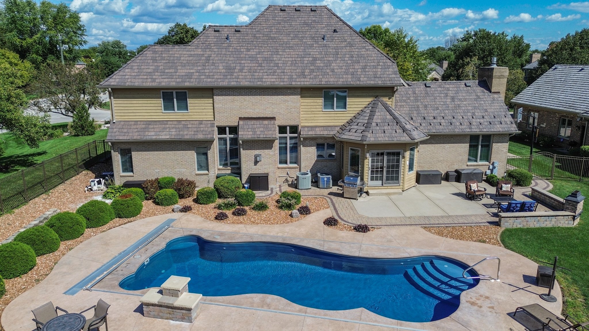 Aerial view of a backyard with a kidney-shaped swimming pool, patio furniture, landscaped bushes, and a large brick house with a tiled roof