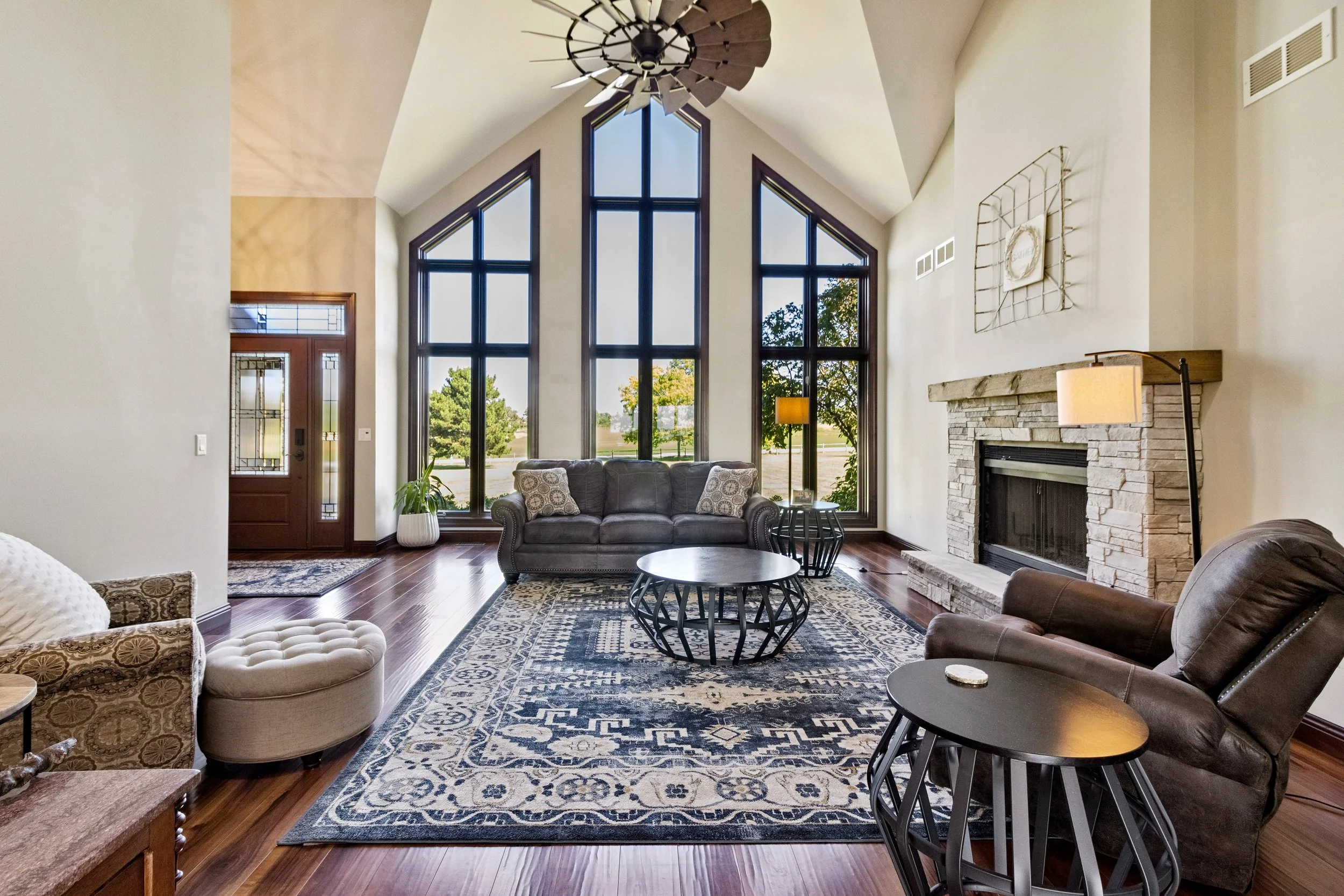 Living room with large front windows, stone fireplace, leather and upholstered furniture, wooden flooring, and a patterned area rug.