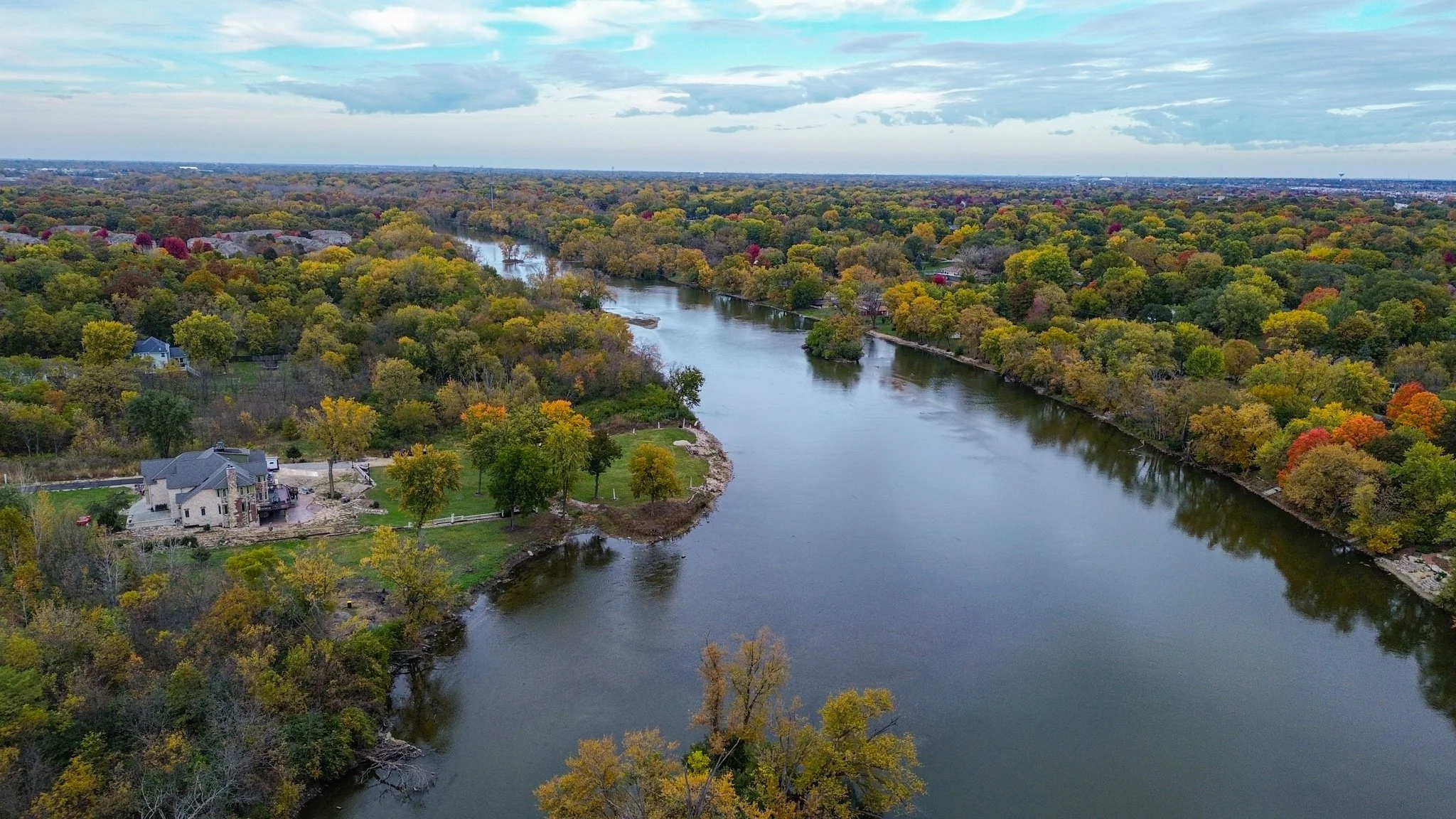 Aerial view of a river winding through a lush, colorful forest with houses along the shoreline, during daytime with partly cloudy sky.