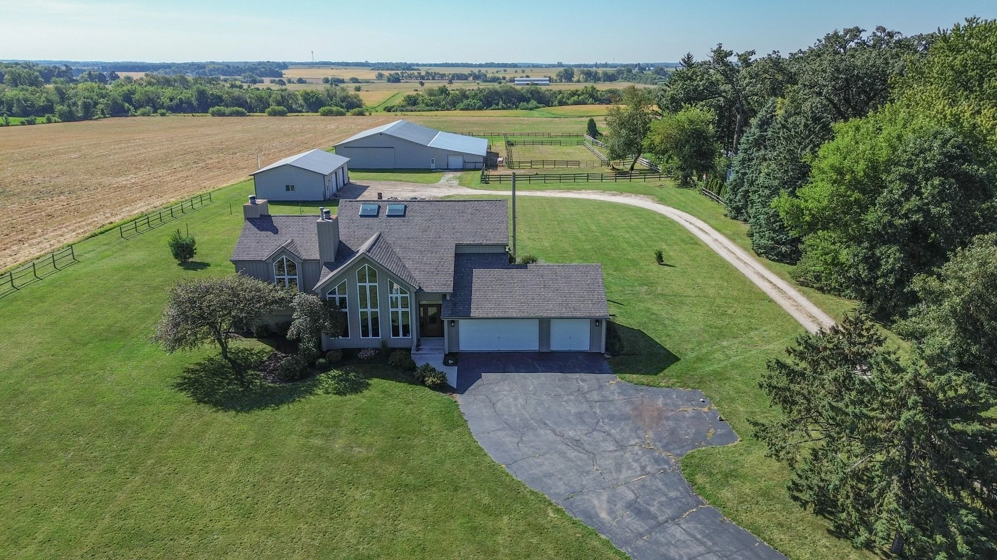 Aerial view of a house with a large lawn, driveway, surrounded by trees and farmland.