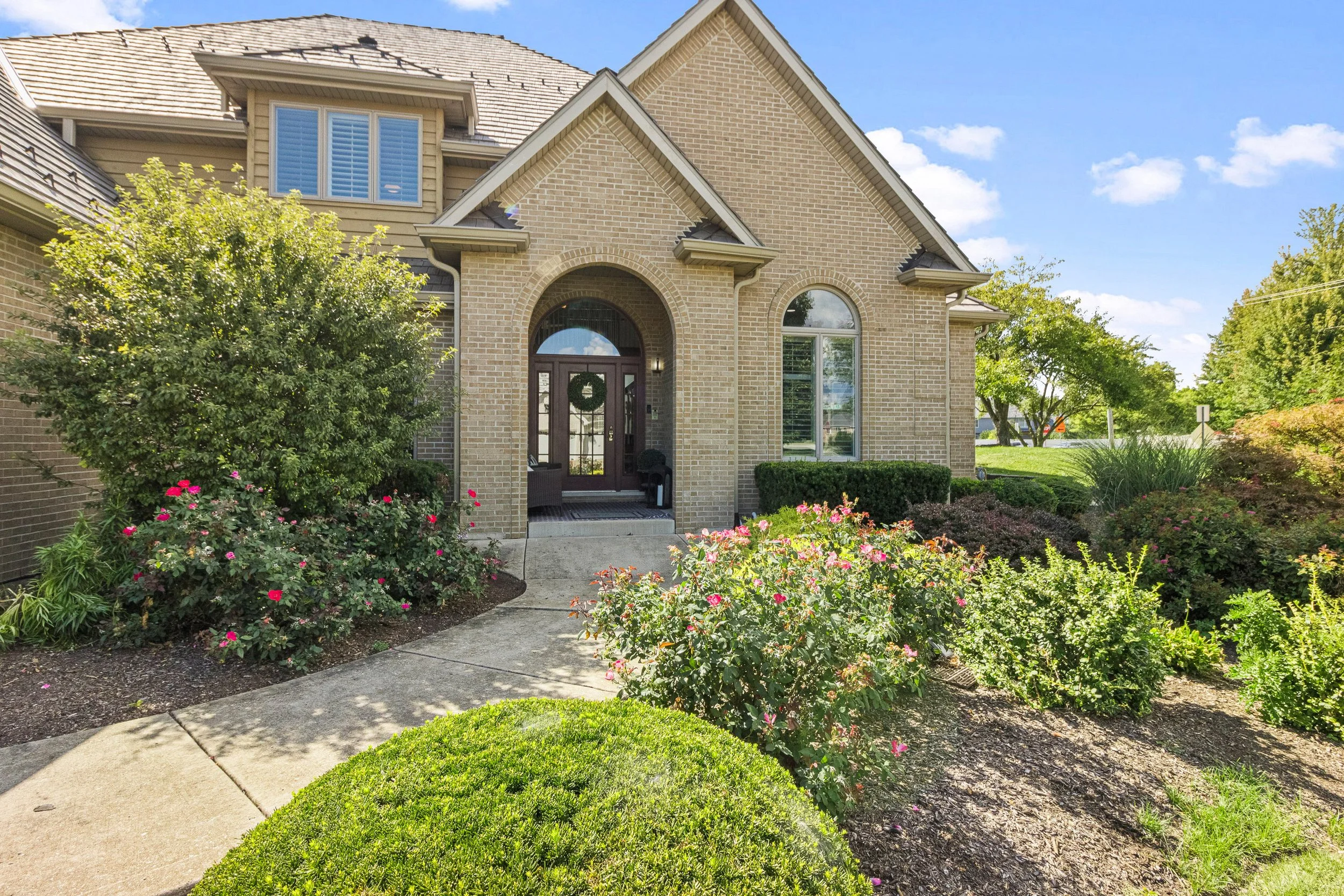 A brick house with a front entrance, surrounded by well-maintained garden bushes and flowers, under a partly cloudy sky.