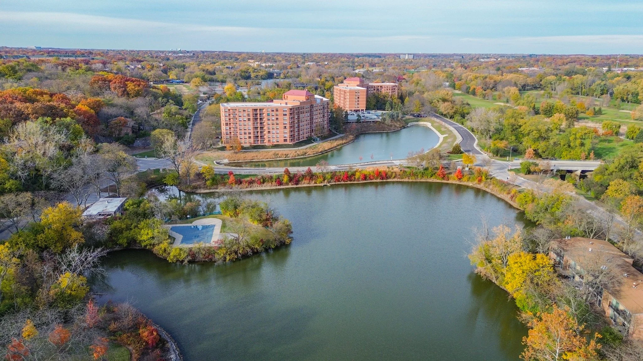 Aerial view of a residential area with trees displaying fall colors, a pond, a smaller pool, and several apartment buildings.