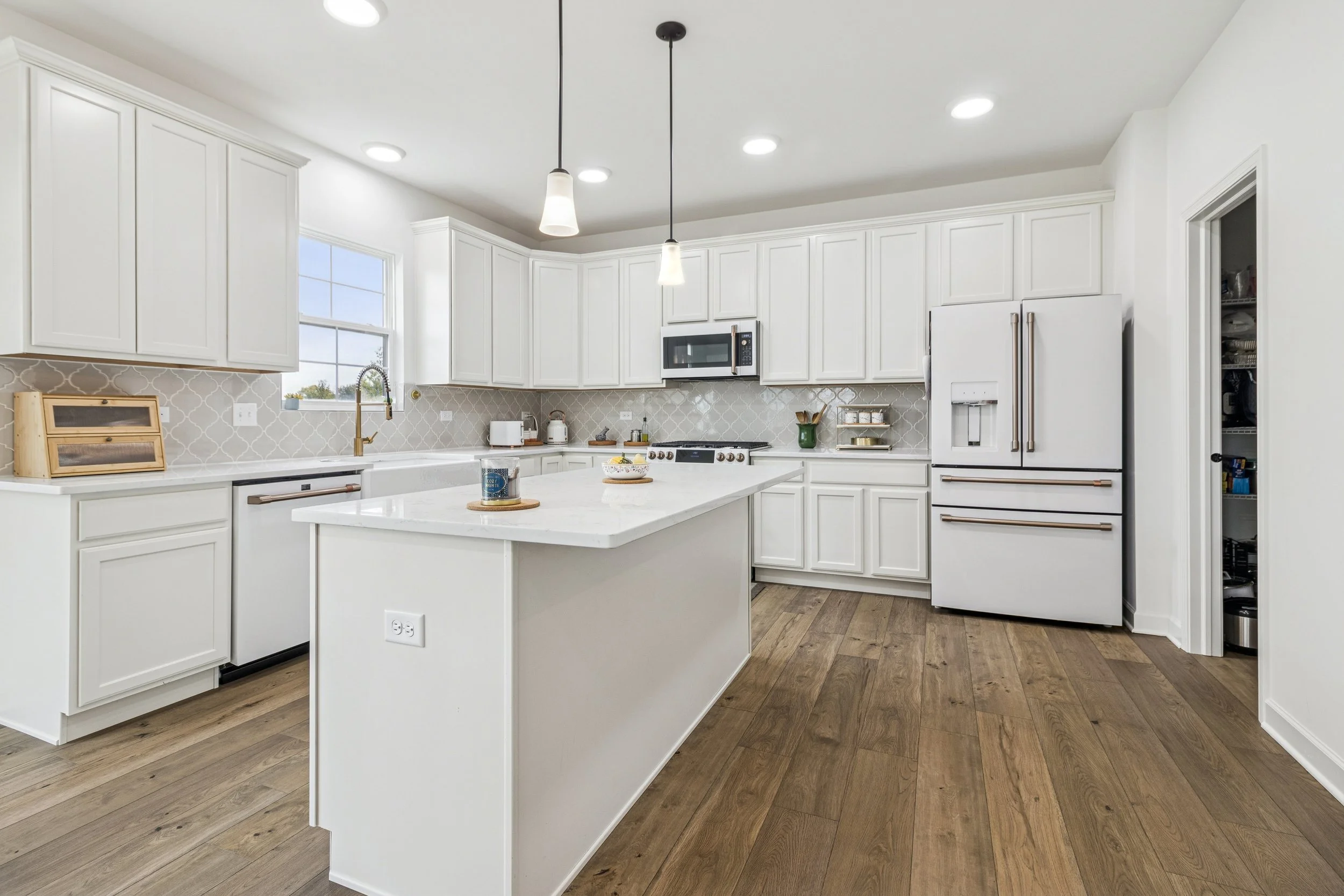 Modern white kitchen with wooden floors, white cabinets, and a central island. Includes appliances like a refrigerator, microwave, and stove. Decorated with a few small kitchen items and pendant lights.