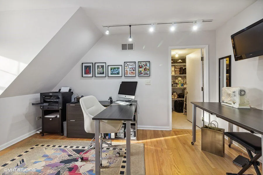 Home office with white walls, hardwood floor, desk, chair, computer, printer, picture frames, wall-mounted TV, and door leading to a walk-in closet.