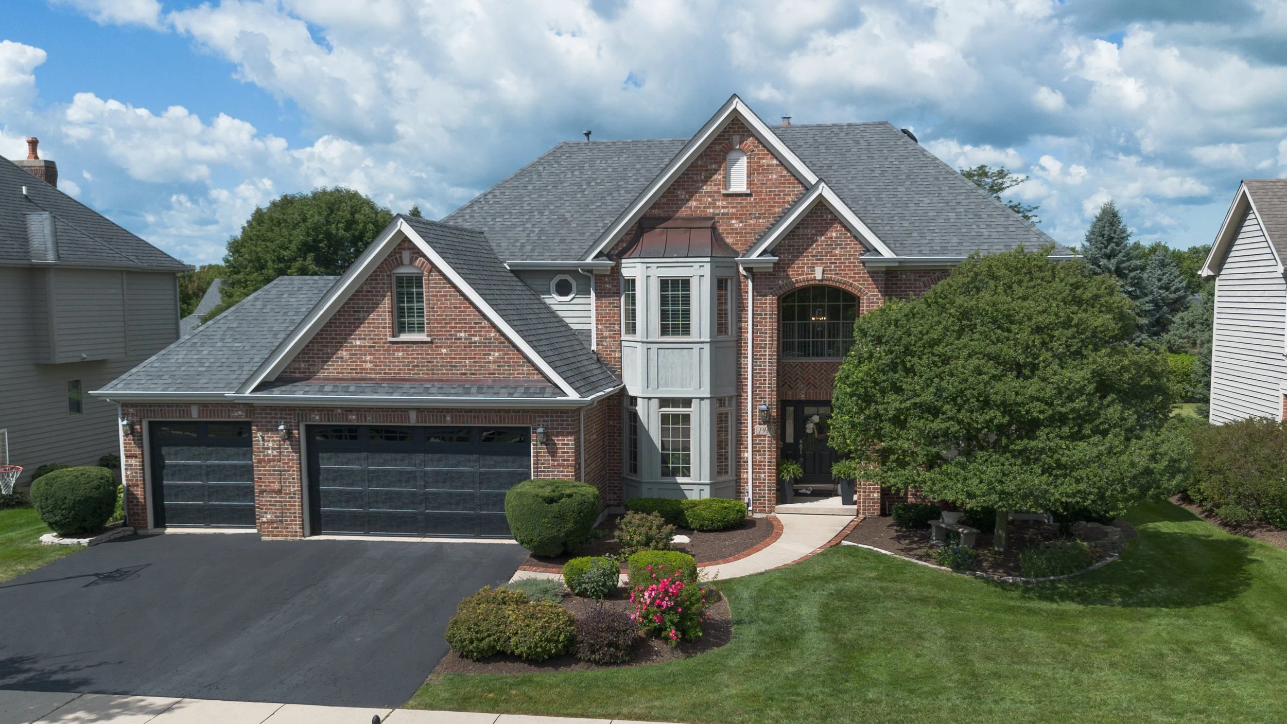 A large two-story brick house with a gray shingled roof, attached garage, a curved pathway leading to the front door, well-maintained lawn, bushes, and a large tree in the front yard. The sky is partly cloudy with blue skies.