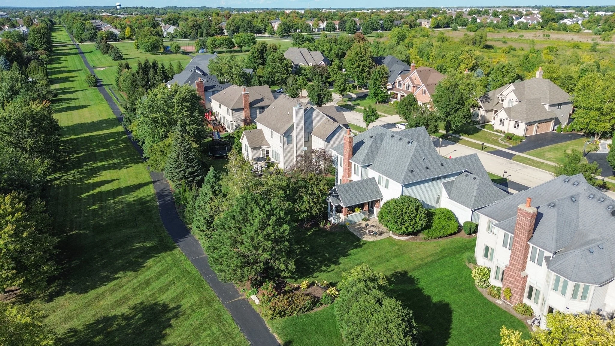 An aerial view of a suburban neighborhood with large houses, trees, and well-manicured lawns, alongside a long green park pathway.