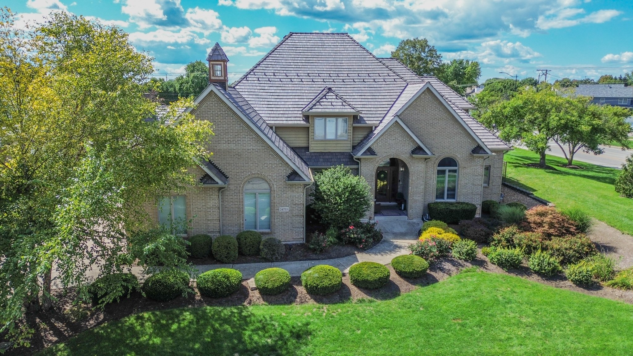A large suburban house with a brick exterior and a steeply pitched roof, surrounded by a well-maintained lawn and landscaped garden with bushes and trees, under a partly cloudy sky.