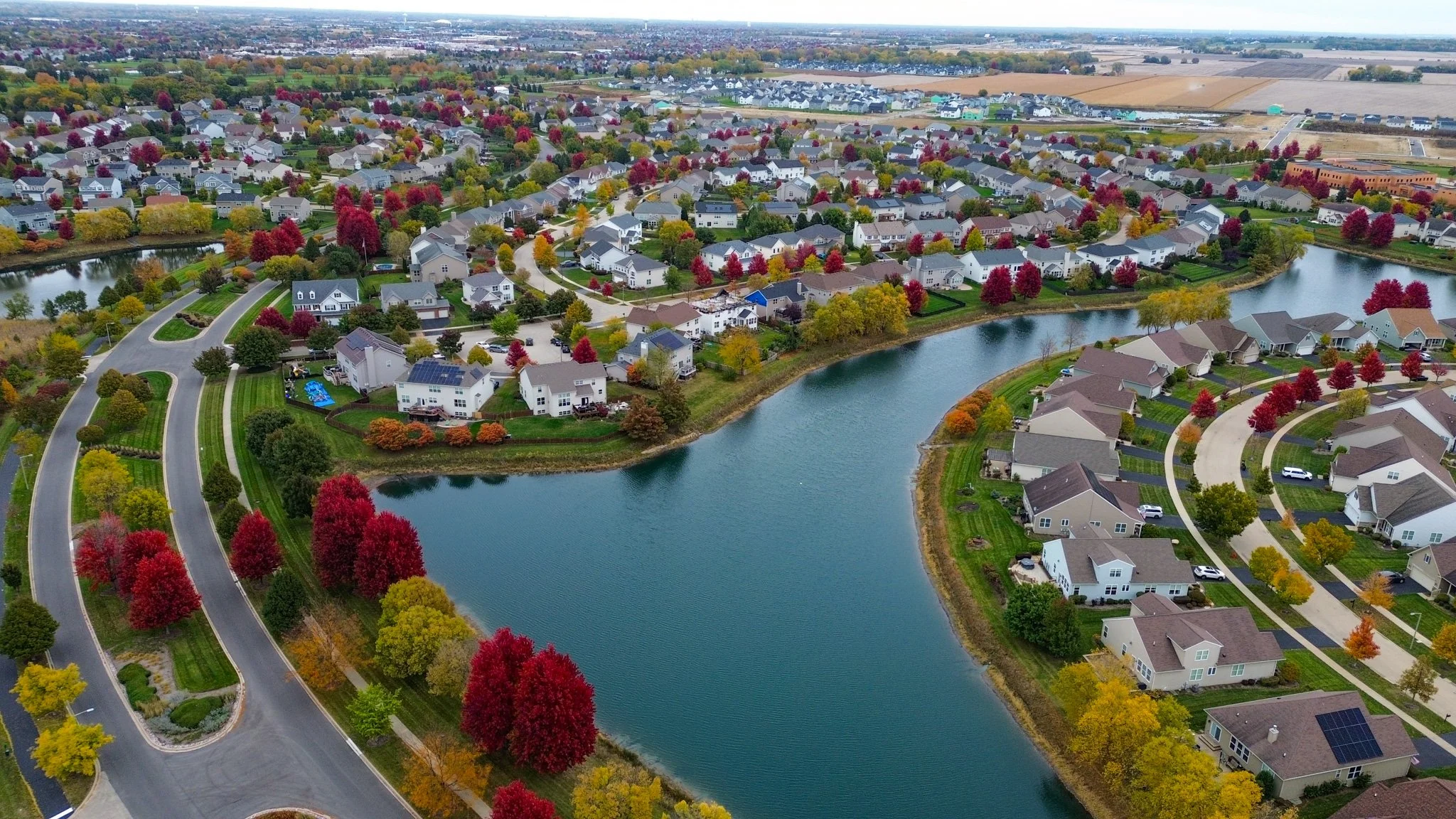 Aerial view of a suburban neighborhood with houses, colorful autumn trees, and a winding lake, showcasing fall foliage.