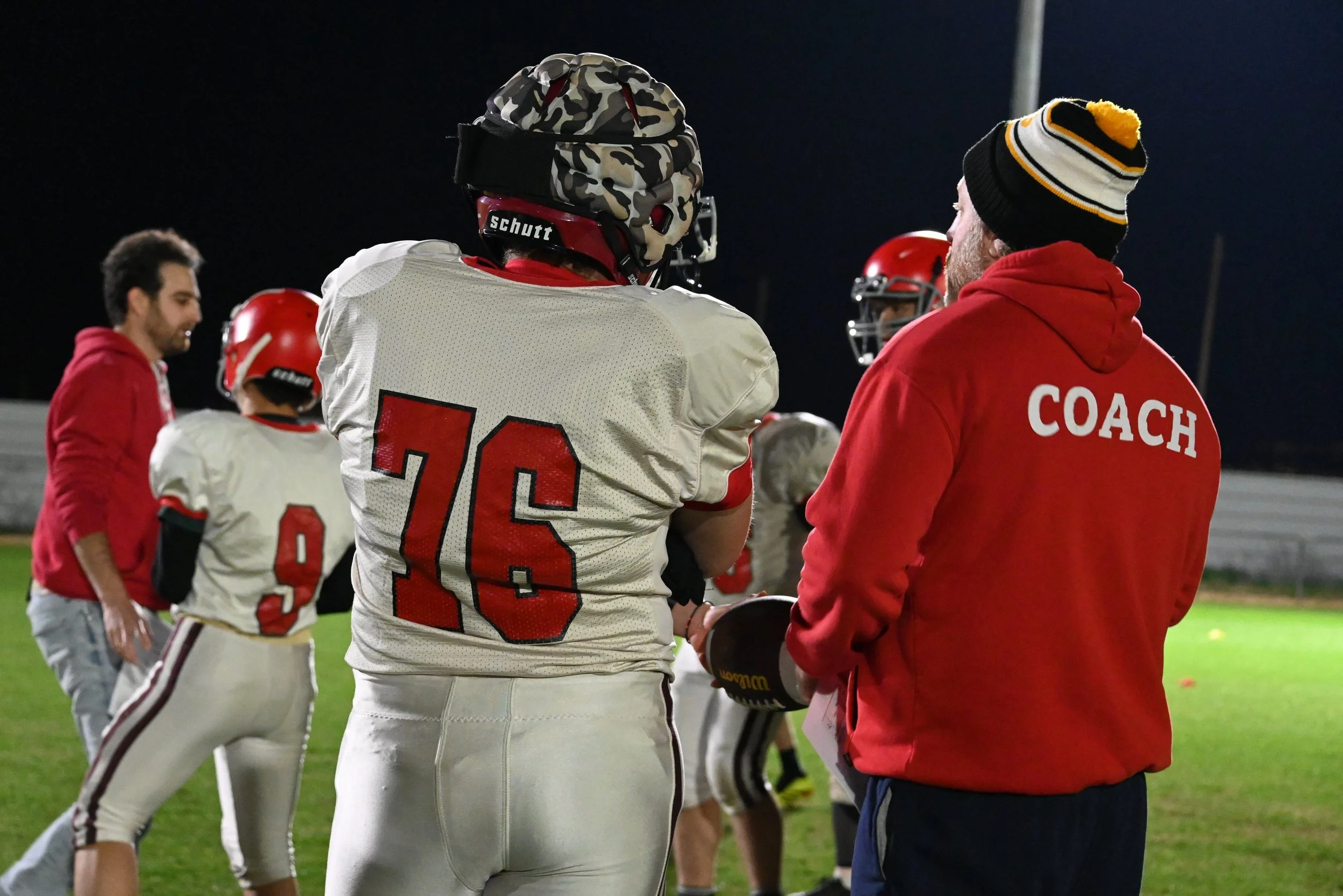 Kadoorie football coach in a red hoodie with 'COACH' printed on the back, talking to a player wearing a white football uniform with the number 76, during a nighttime game or practice on the field, with other players and staff in the background.