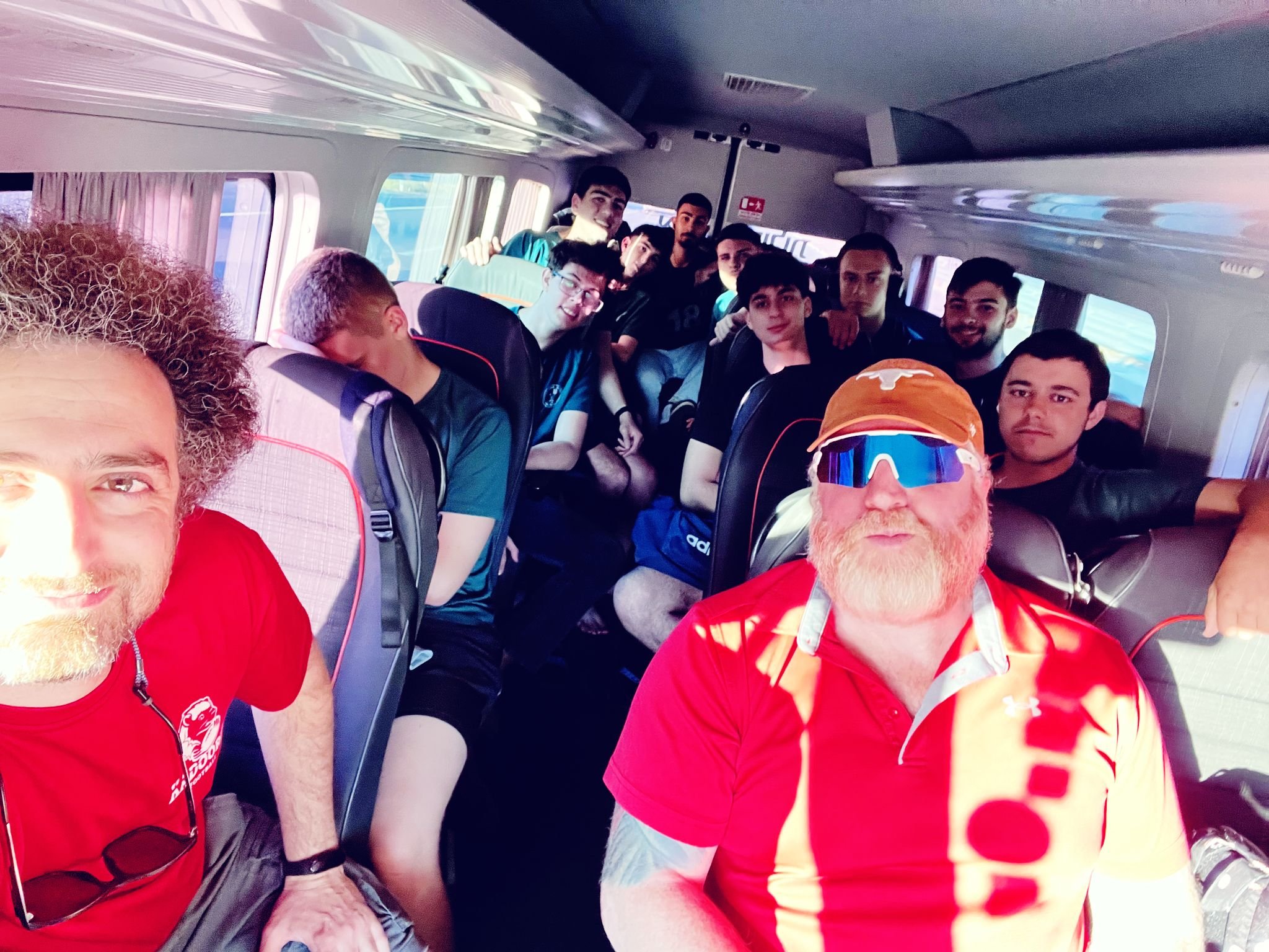 Kadoorie football team sitting inside a bus, with coaches in the foreground wearing red shirts. Players are seated in the background, some smiling and looking at the camera.