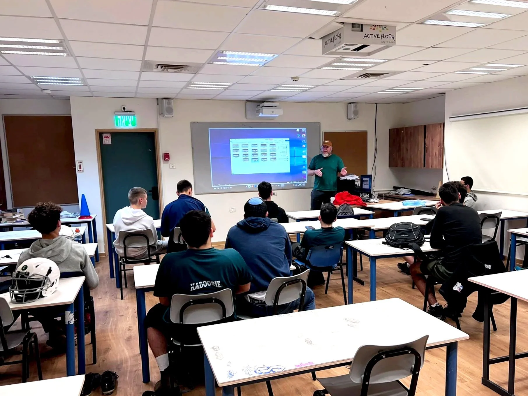Kadoorie football players attending a classroom lecture, seated at desks, facing a coach who is standing next to a large interactive whiteboard display showing a computer screen.