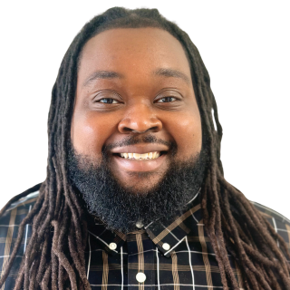 A man with long dreadlocks and a beard, smiling, wearing a dark plaid shirt.