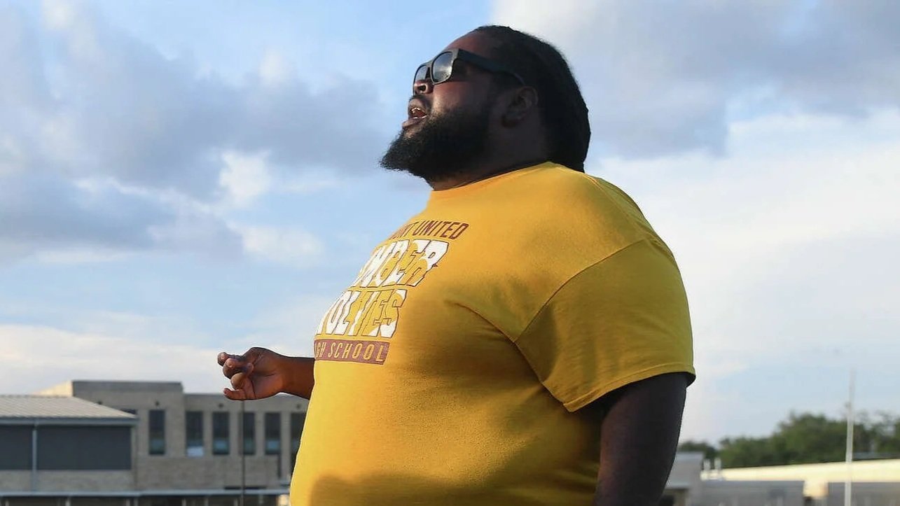 A man with a beard and sunglasses wearing a yellow T-shirt standing outside with a partially cloudy sky in the background.
