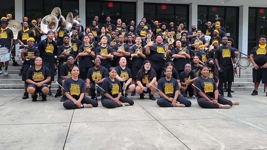 A large group of marching band members in black shirts with yellow text, posing on steps outside a building, some holding musical instruments such as drums and brass instruments.