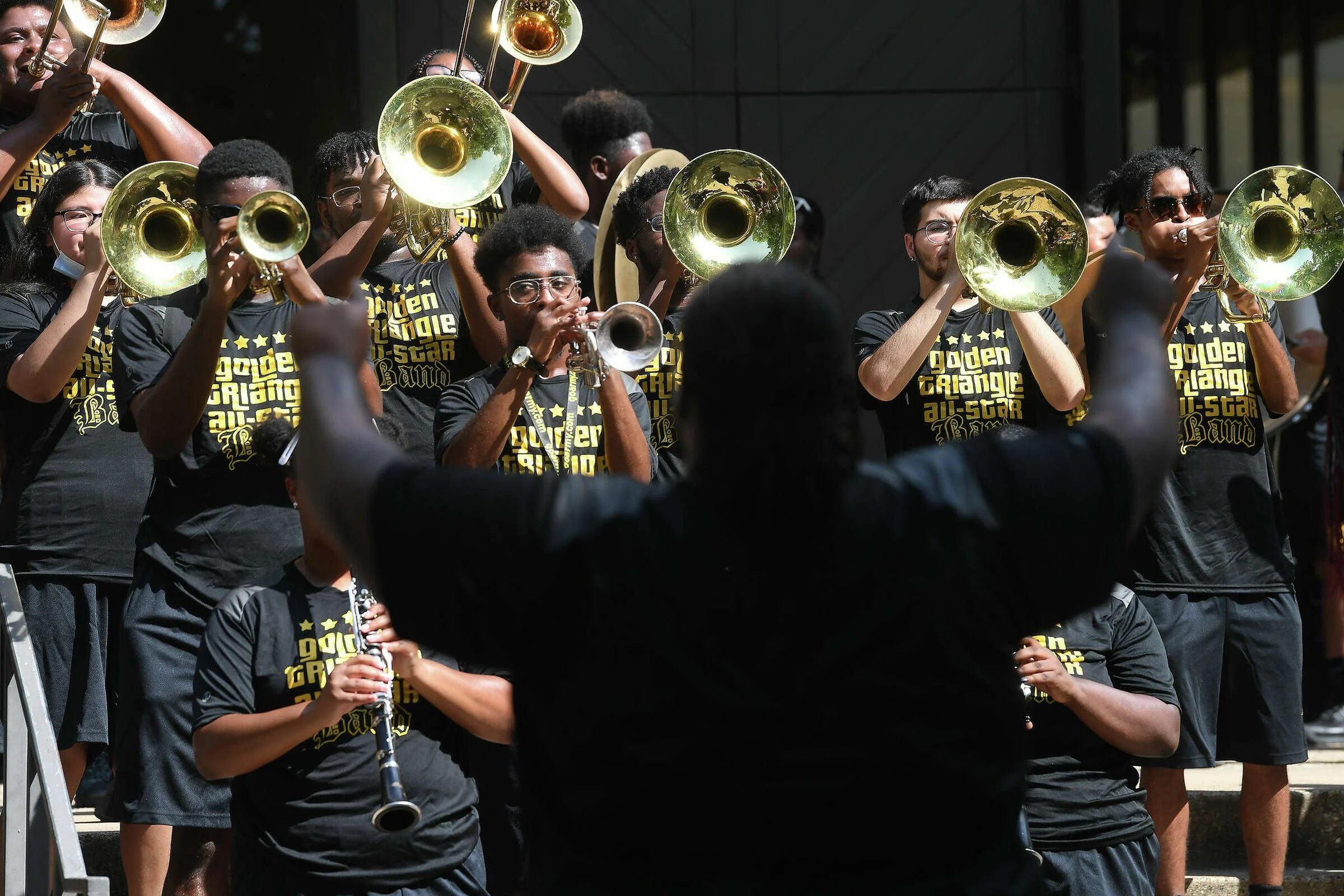 A group of young musicians playing brass instruments, directed by a conductor, outdoors during daytime. The musicians wear black shirts with yellow text, some have glasses, and they appear to be part of a band performing together.