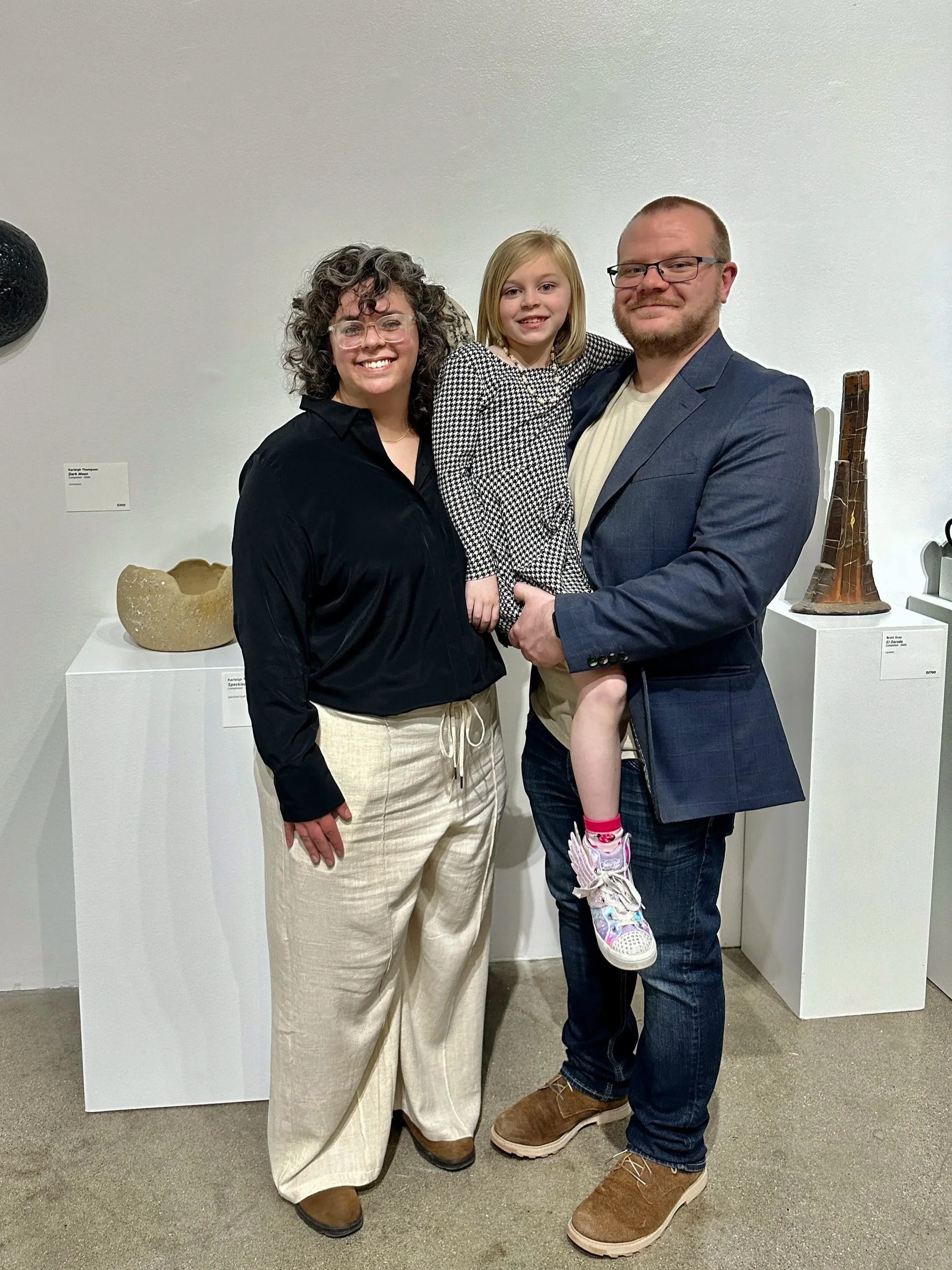 A woman with curly hair and glasses, a young girl, and a man with glasses are standing together in an art gallery, smiling at the camera. The man is holding the girl, who is wearing a checkered dress and colorful sneakers. They are in front of white pedestals with ceramic sculptures.