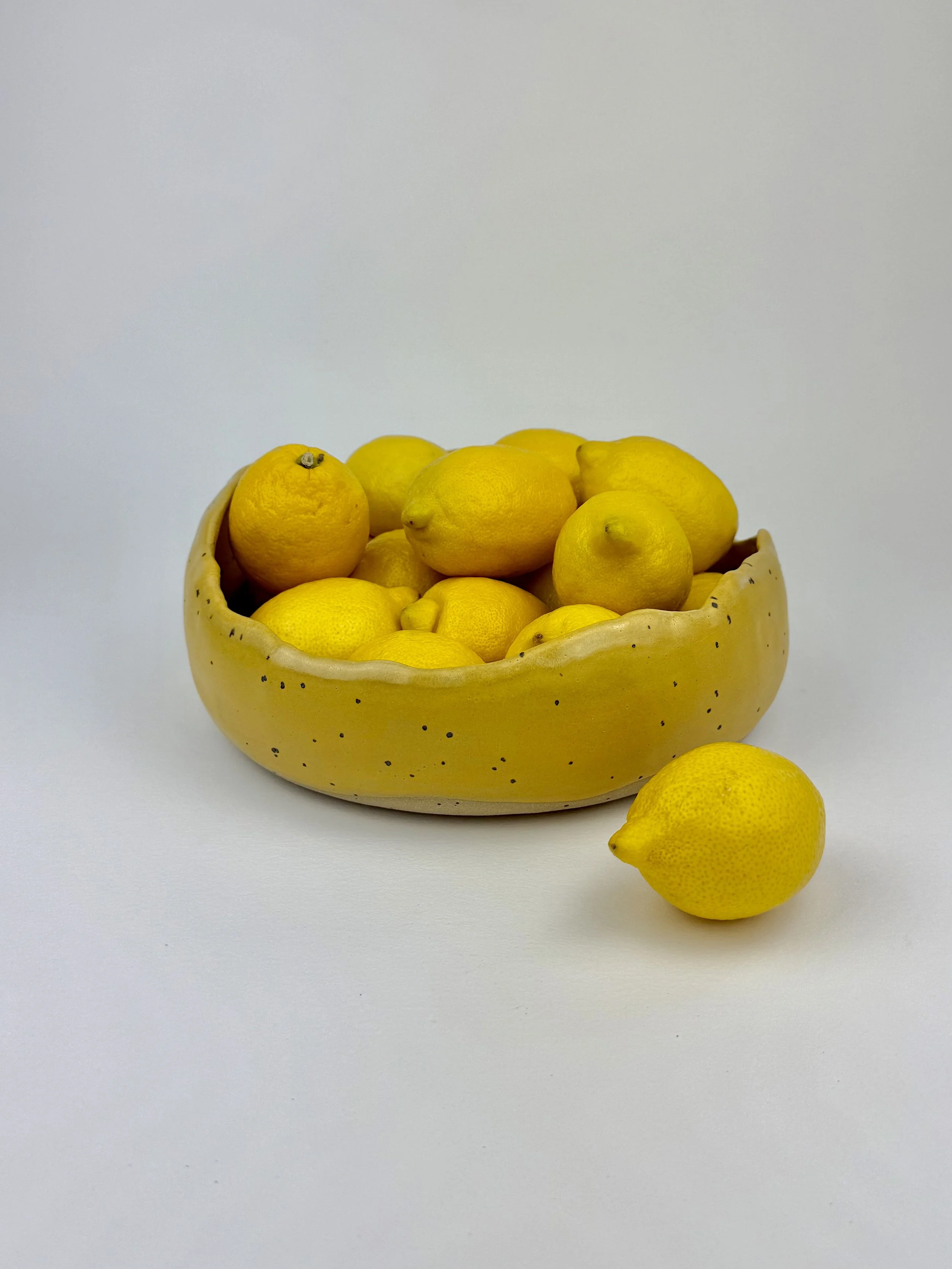 Ceramic bowl filled with yellow lemons, with one lemon placed outside the bowl on a white background.