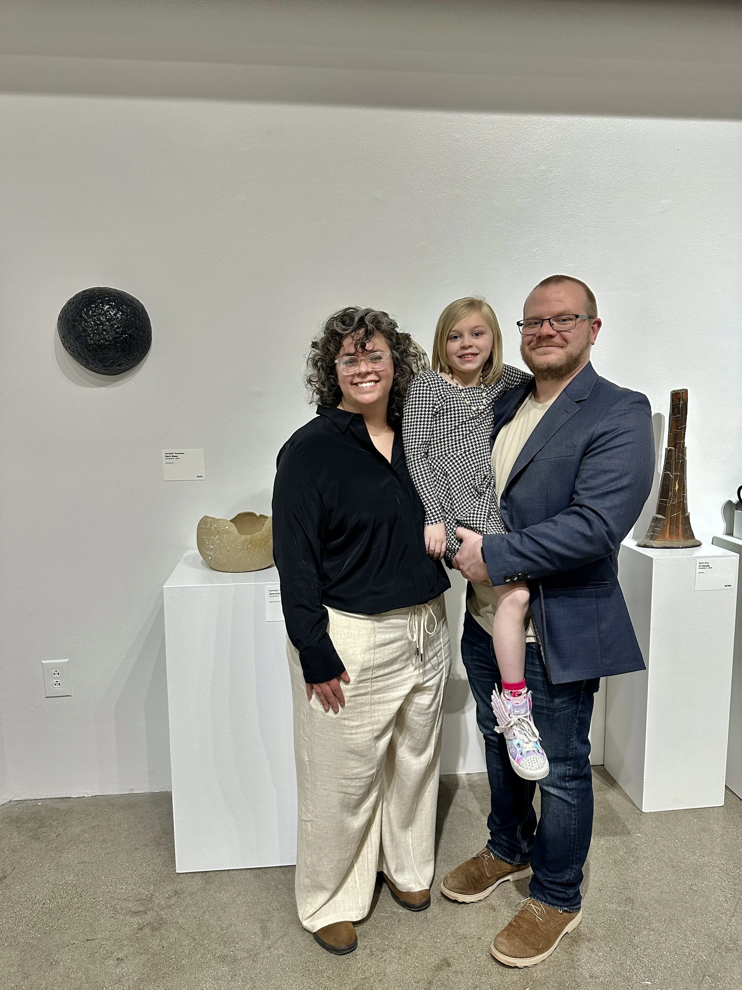 A family of three posing together in an art gallery, with artwork displayed on the wall behind them.