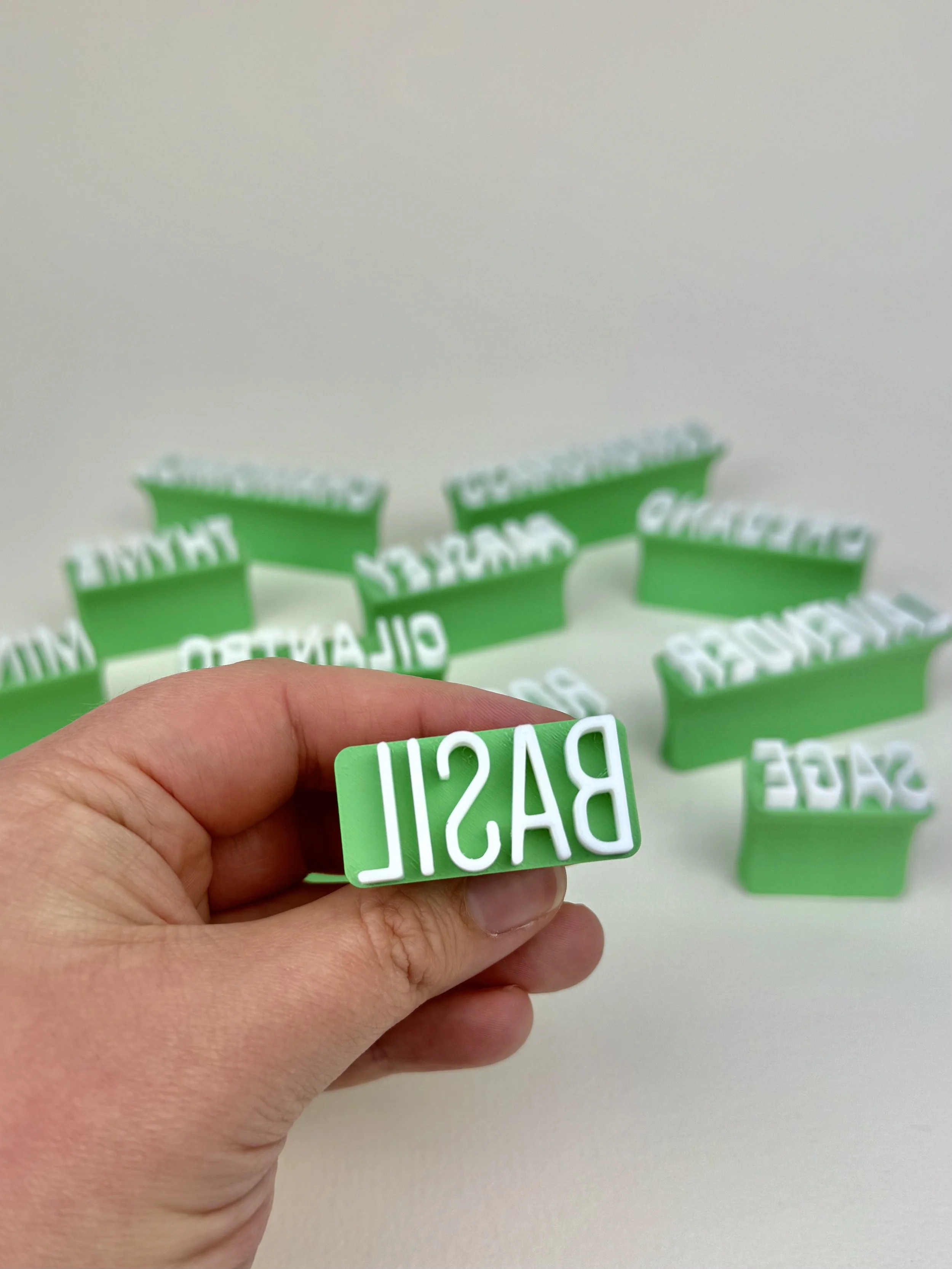 Hand holding a green stamp with the word "BASIL" in white, with multiple similar stamps scattered on a white surface in the background.