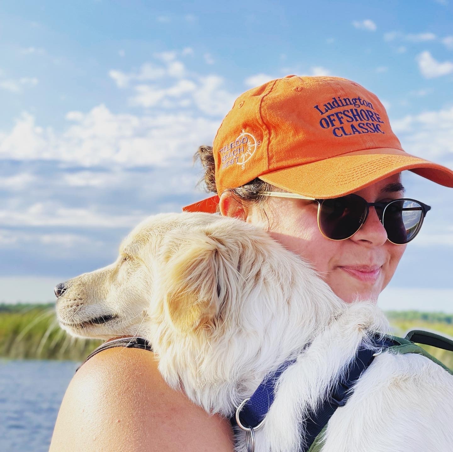 A woman wearing sunglasses and an orange cap with text, holding a white dog with long ears, in an outdoor setting with water and greenery in the background.