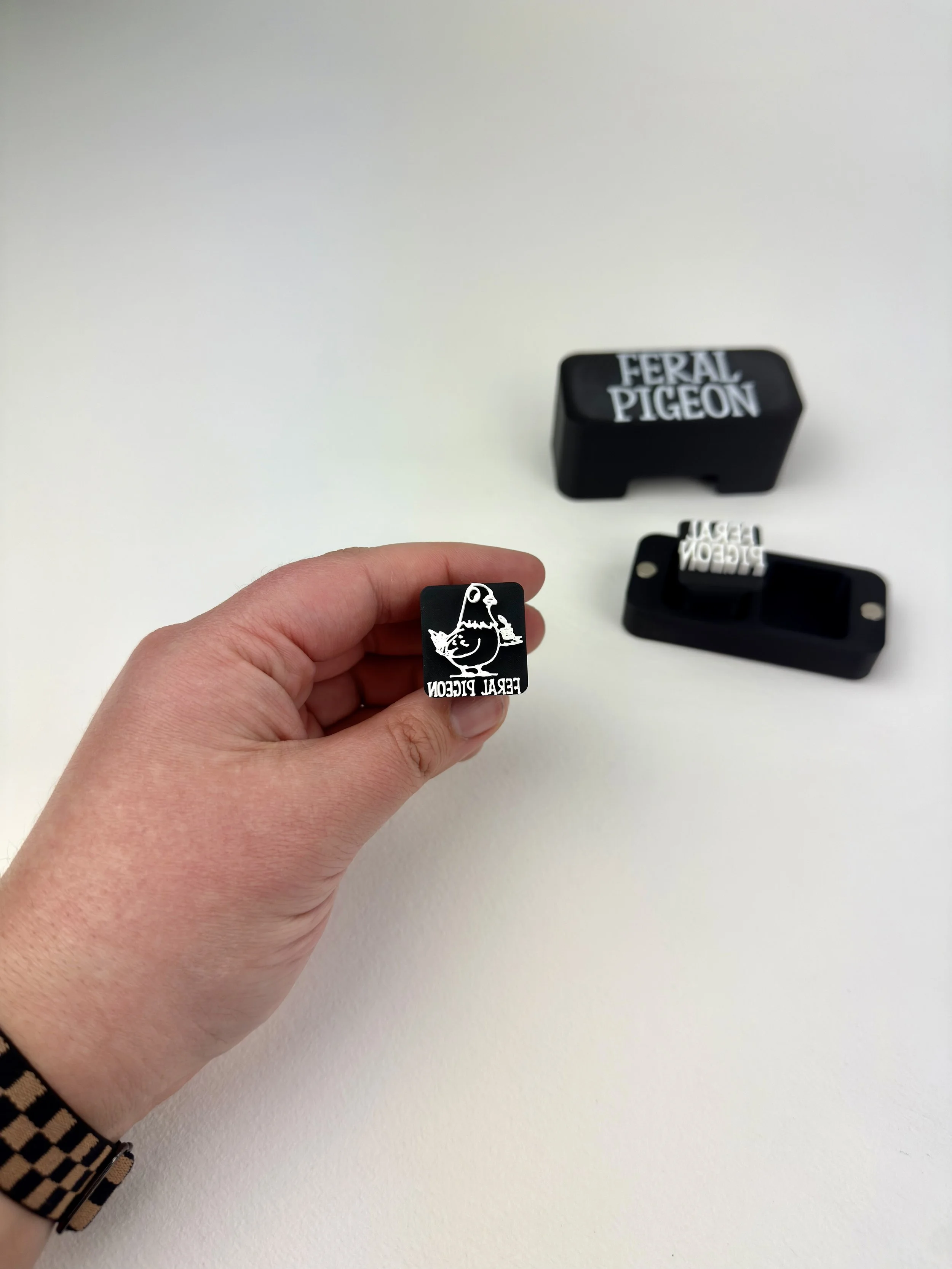 Hand holding a black rubber stamp with a duck illustration and the text 'FERAL PIGEON'. In the background, there are two more stamps and a black case with the text 'FERAL PIGEON'.