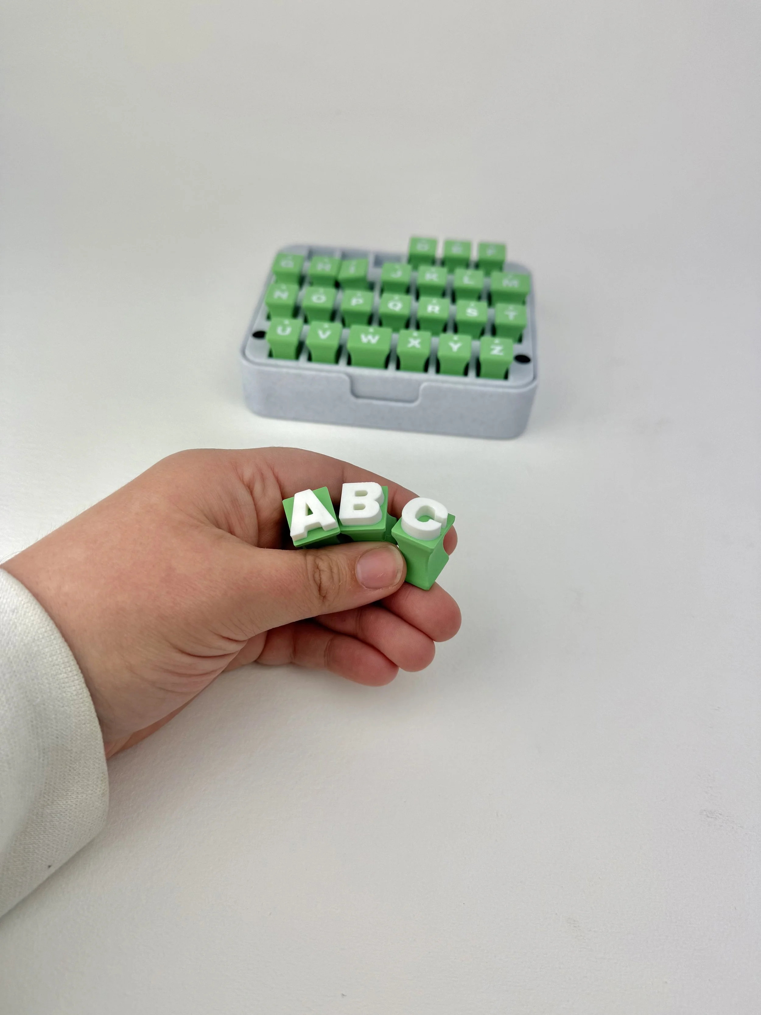 A hand holding three small green blocks with white letters spelling 'ABC'. In the background, there is a box of similar blocks with letters on them, sitting on a white surface.