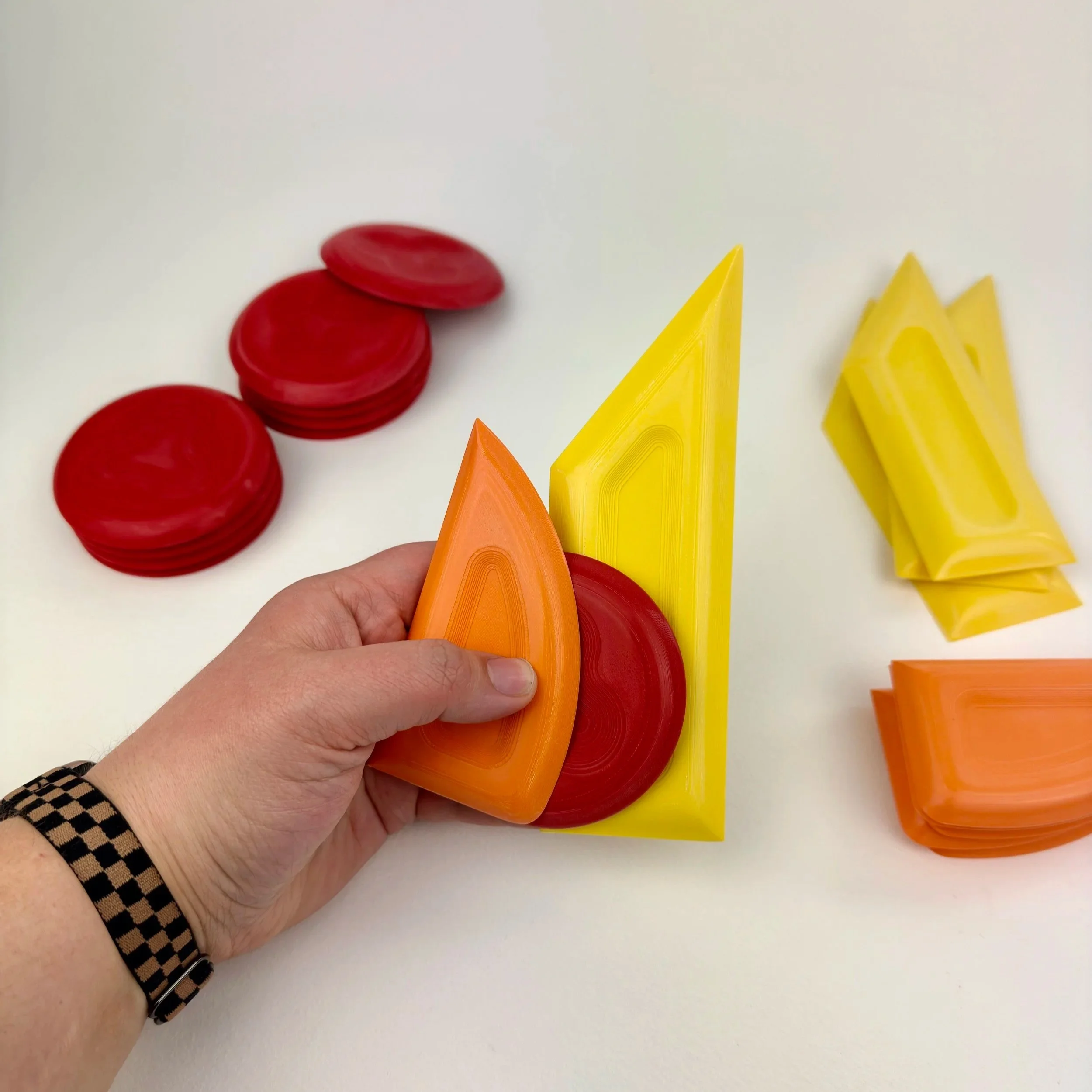 Person holds an orange and yellow triangular and semi-circular acrylic pieces, with other red and yellow acrylic pieces on a white surface.